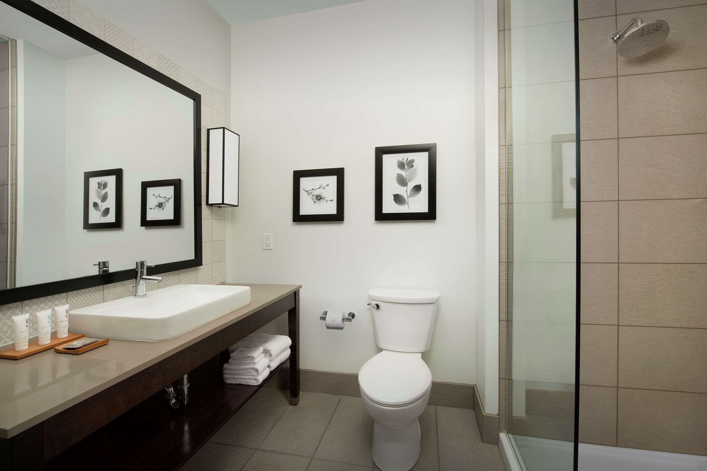 A modern bathroom with a large mirror, white sink, and toilet. There are framed pictures on the wall and a glass shower door.