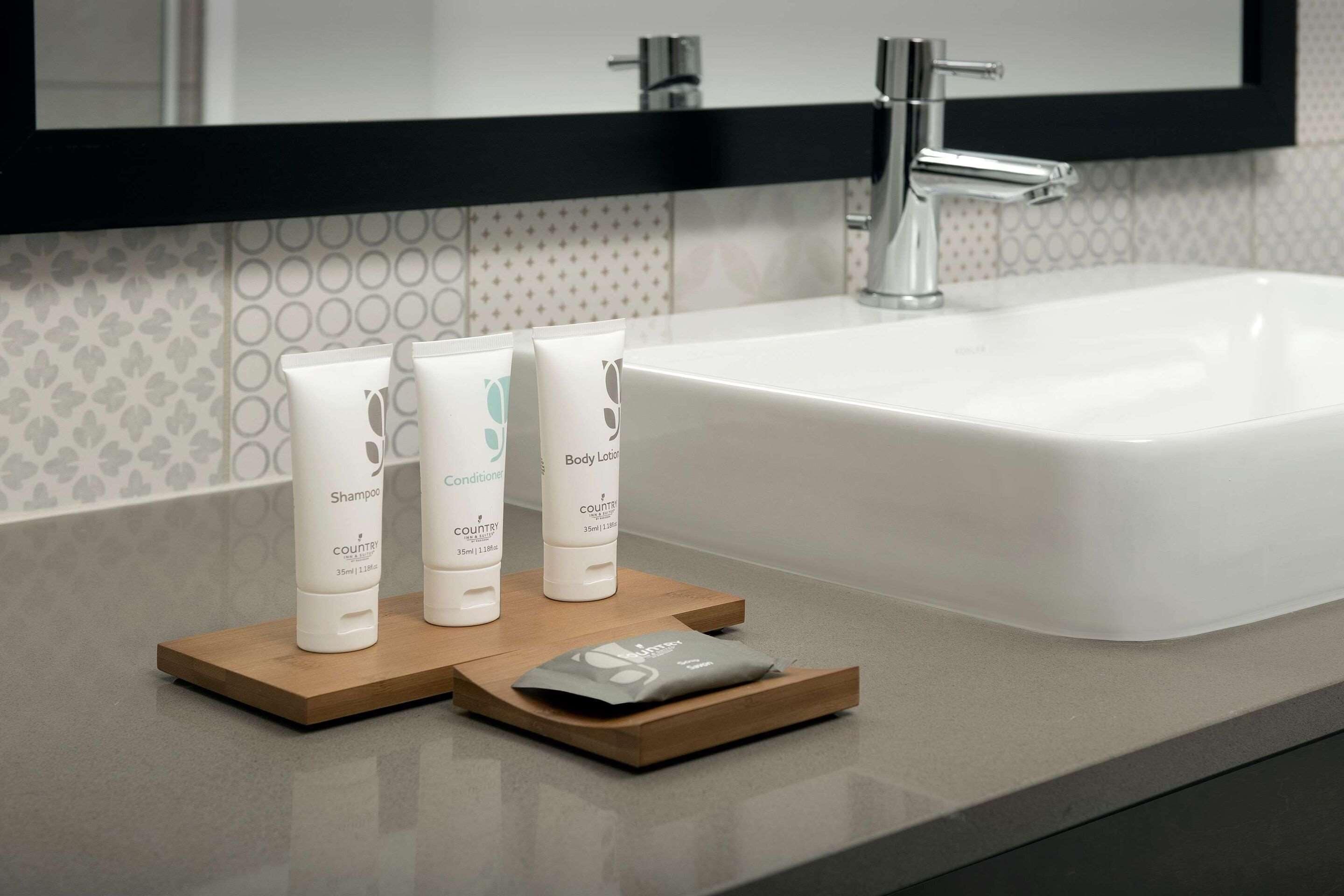 Une salle de bain avec un lavabo blanc, deux flacons de produits de toilette blancs posés sur un plateau en bois, et un miroir au-dessus du lavabo.