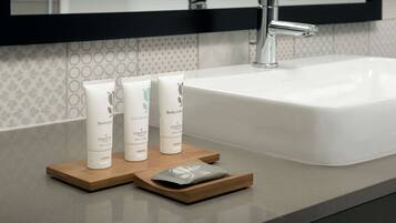 Une salle de bain avec un lavabo blanc, deux flacons de produits de toilette blancs posés sur un plateau en bois, et un miroir au-dessus du lavabo.