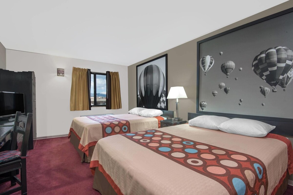 A hotel room with two beds, a desk with a chair, a TV, a window with curtains, a lamp, and a wall with hot air balloon artwork.