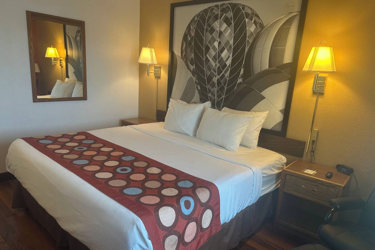 Une chambre d’hôtel avec un grand lit, deux lampes de chevet, une table de chevet, une chaise et un grand tableau abstrait accroché au mur.