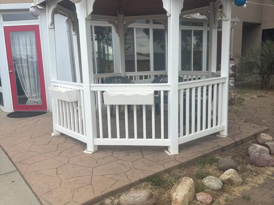 A gazebo with a white railing and a red roof, situated on a paved area with a stone border.