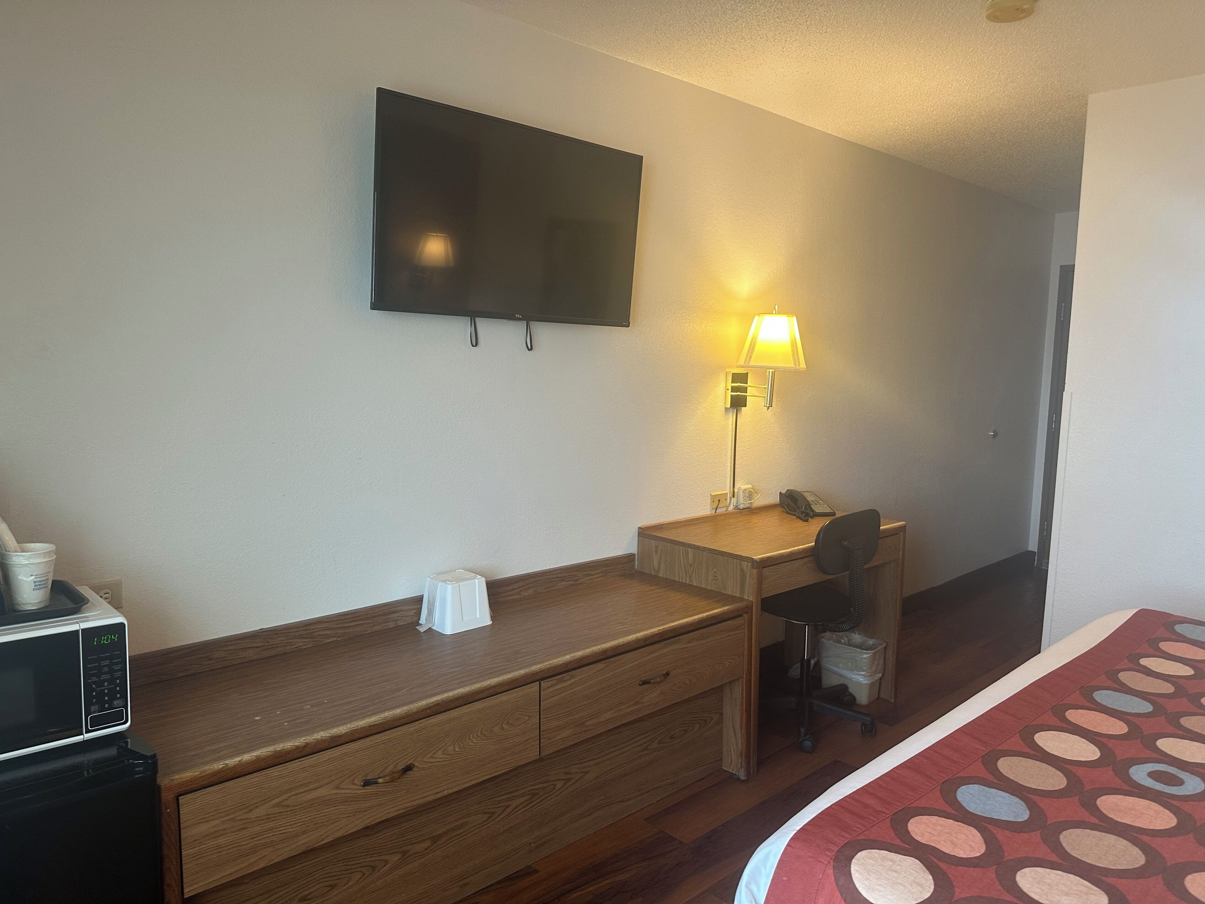 A hotel room with a wooden desk, a chair, a television mounted on the wall, a lamp, a microwave, and a bed with a patterned bedspread.