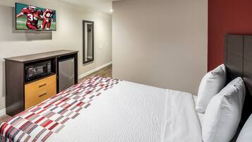 A hotel room with a bed, a TV mounted on the wall, a small desk with a refrigerator, and a mirror.