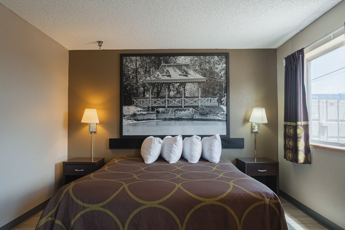 Een hotelkamer met een bed, twee nachtkastjes, twee lampen, een groot gepresenteerd schilderij aan de muur en een raam met gordijnen.