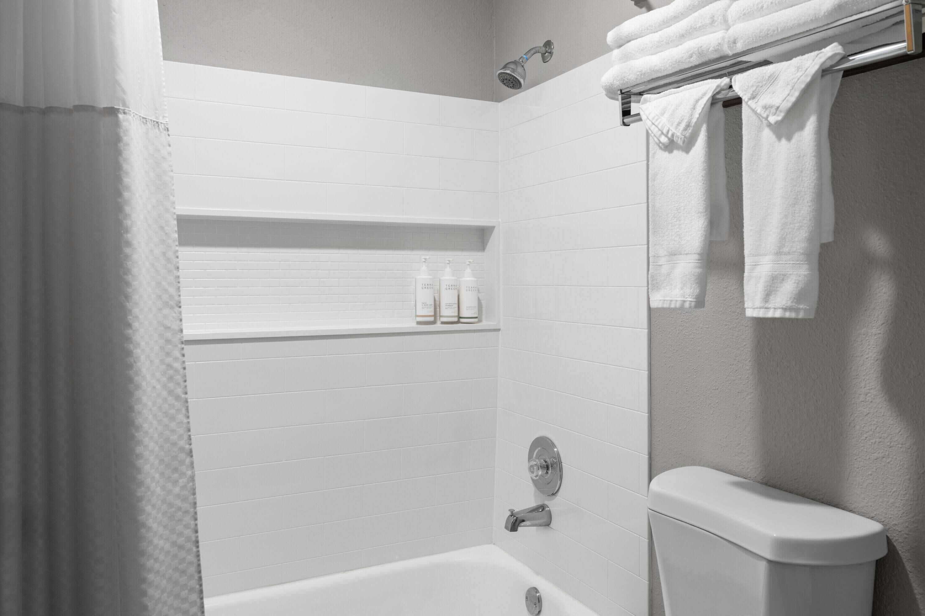 A modern bathroom with a bathtub, a toilet, and a shower. There are white towels hanging on a rack and a shelf with toiletries.