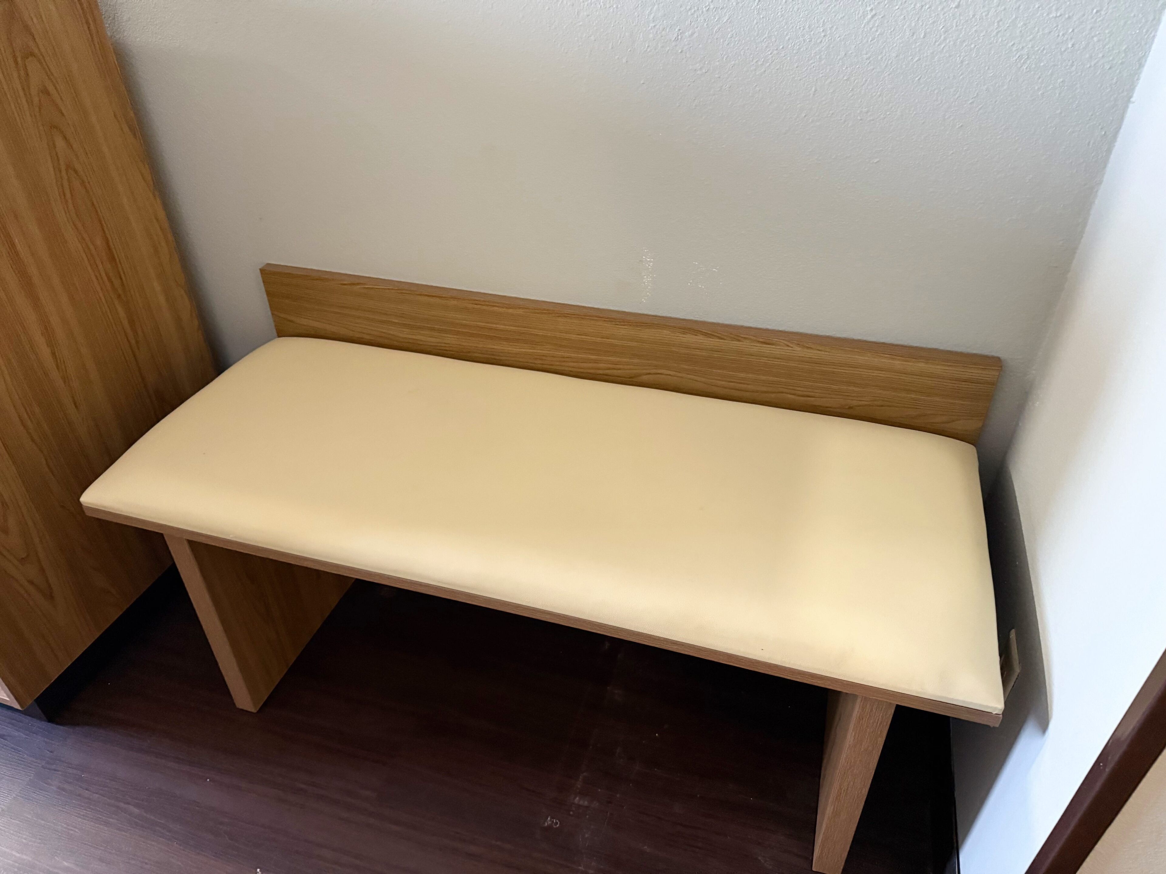 A small, wooden bench with a light-colored cushion in a corner of a room with dark wooden flooring and white walls.
