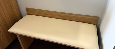 A small, wooden bench with a light-colored cushion in a corner of a room with dark wooden flooring and white walls.