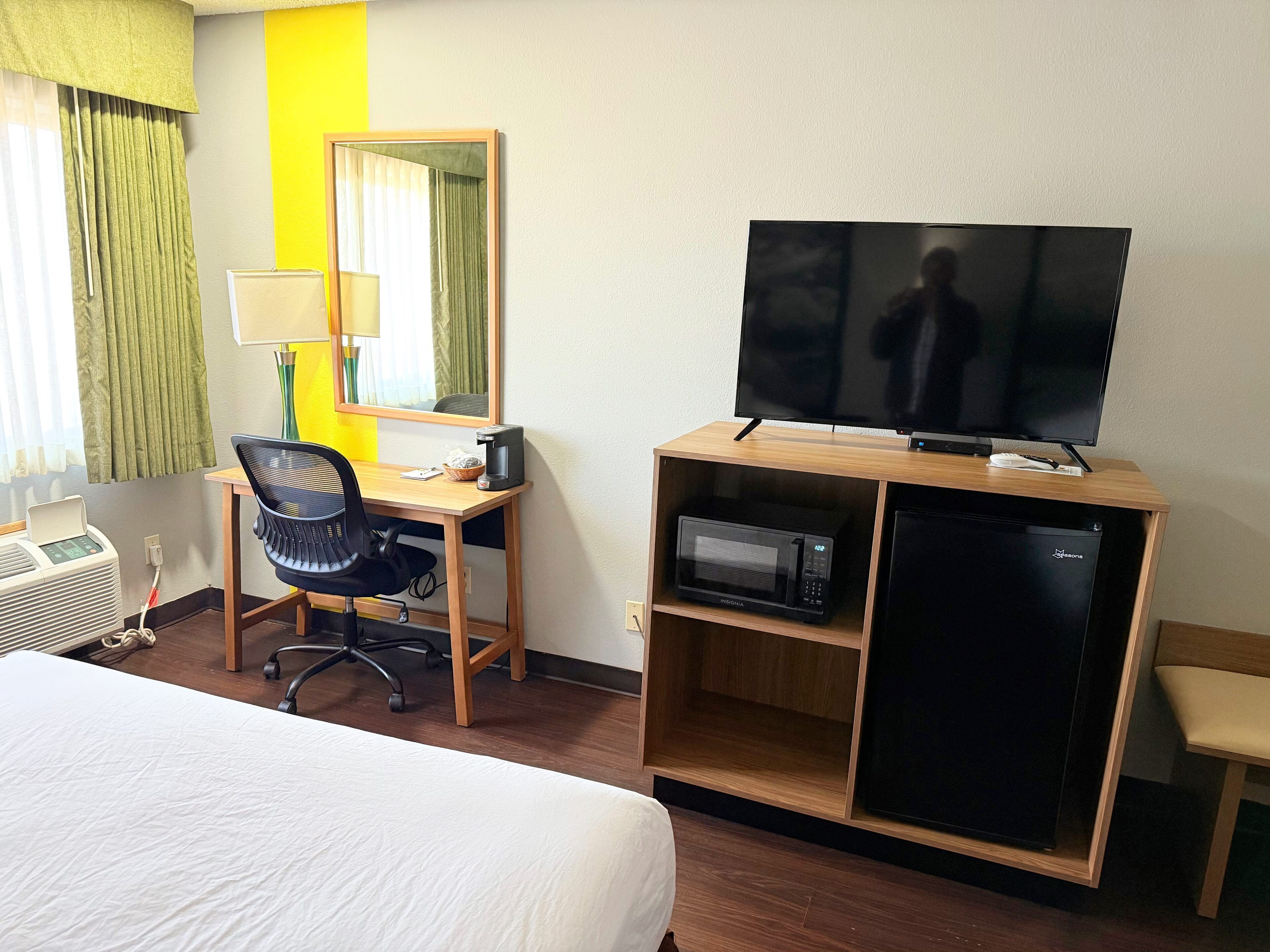 Una habitación de hotel con una cama, un escritorio con una silla, un televisor, un microondas y un refrigerador pequeño.
