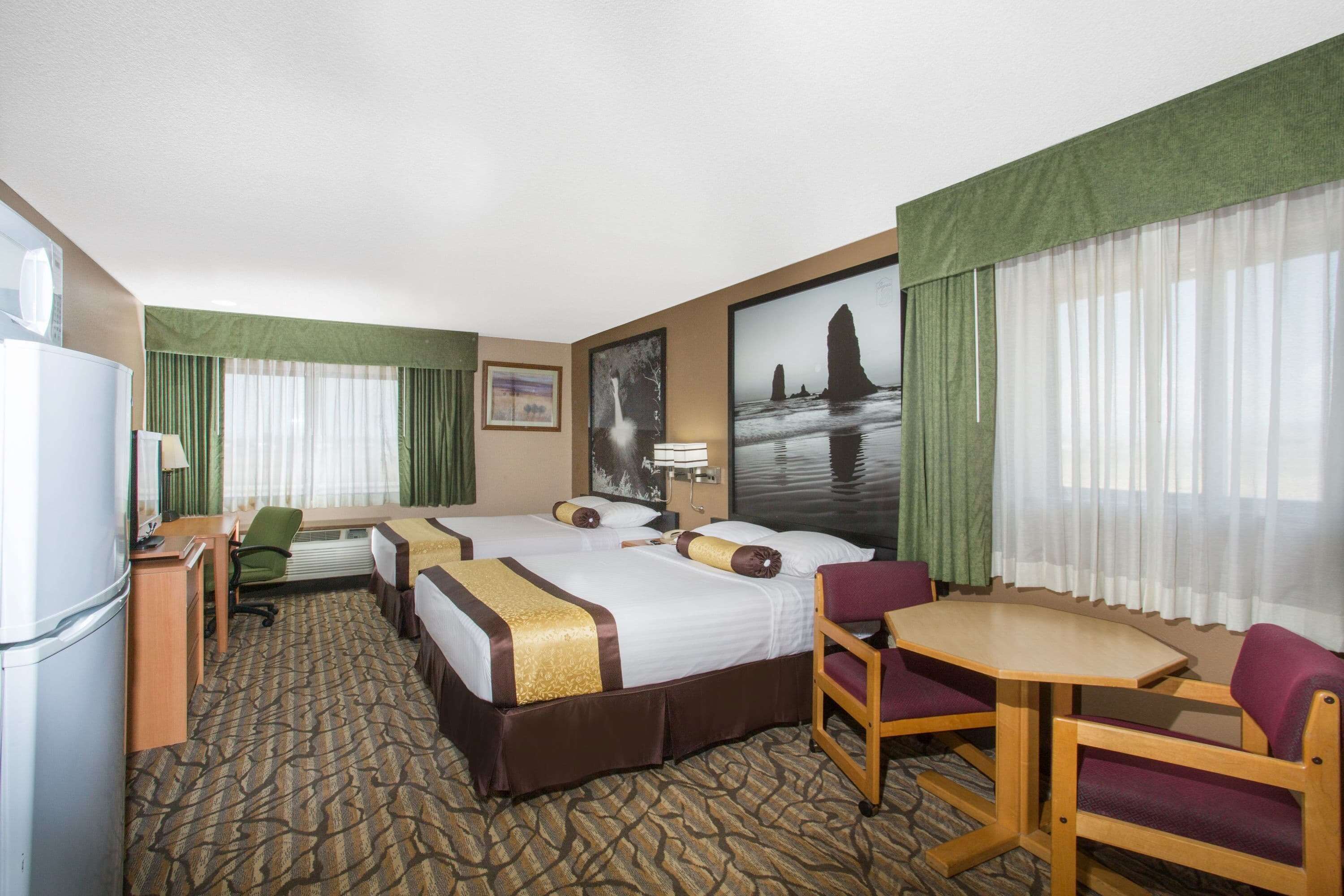 A hotel room with two beds, a desk, a chair, a small table, and a refrigerator.