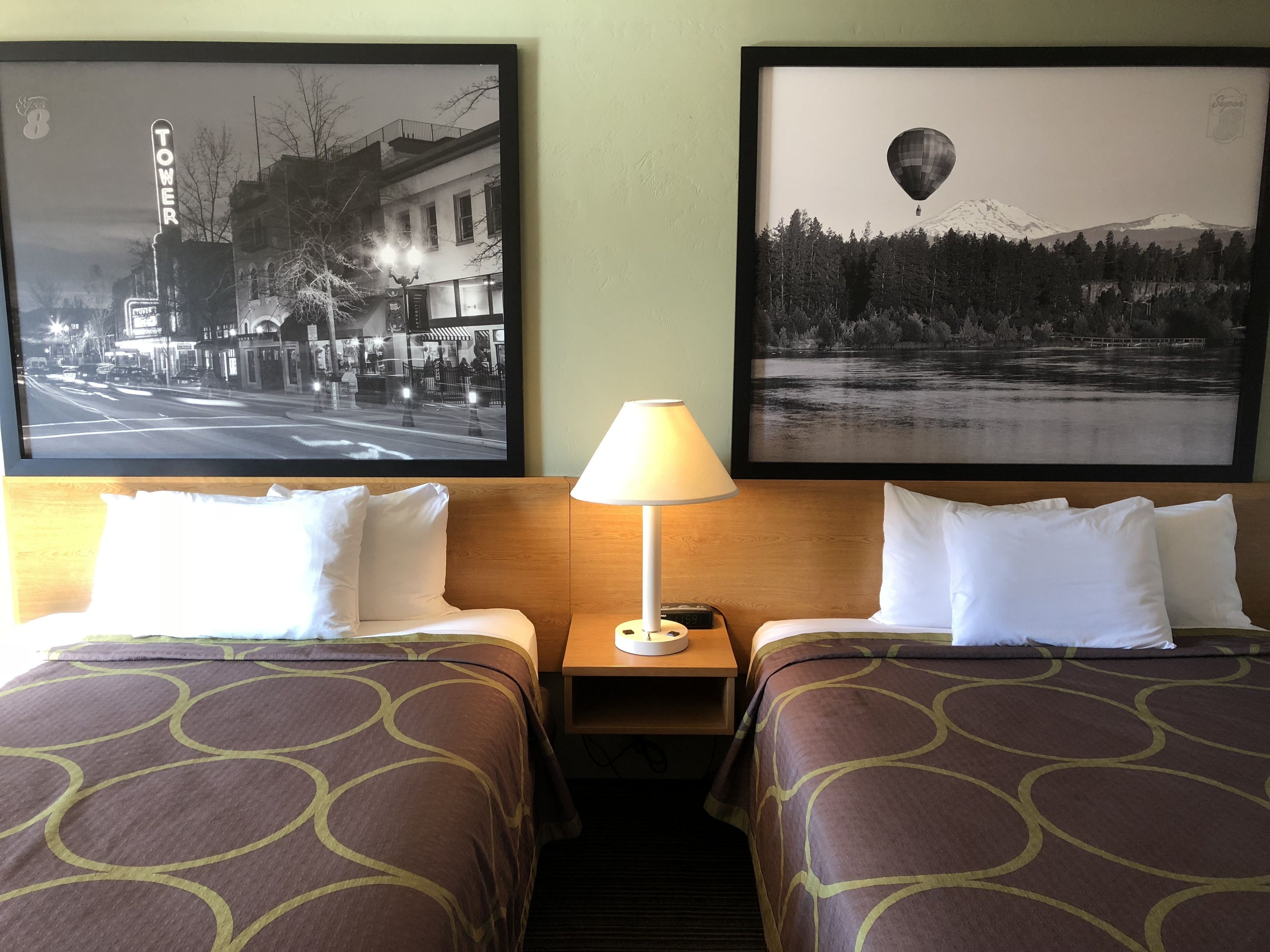 A hotel room with two beds, a nightstand with a lamp, and two framed pictures on the wall.