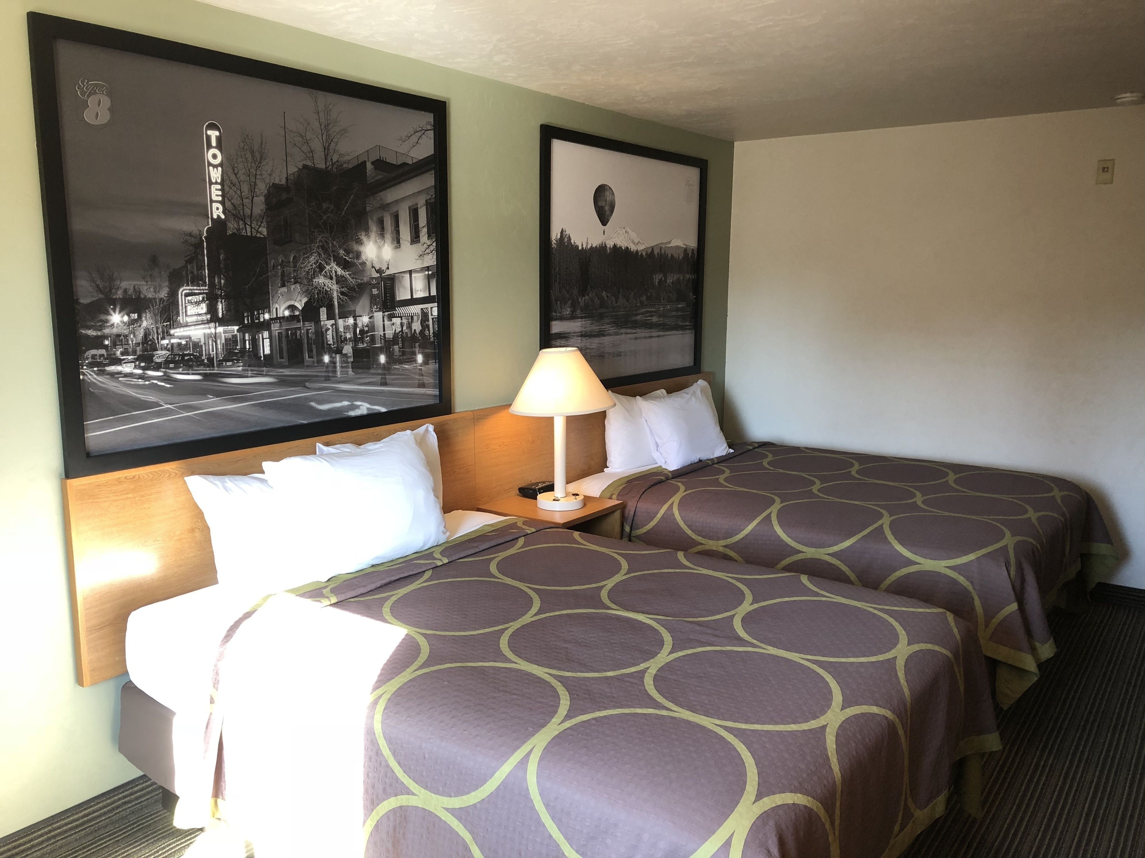 Habitación de hotel con dos camas, una mesita de noche, una lámpara y dos cuadros en la pared.