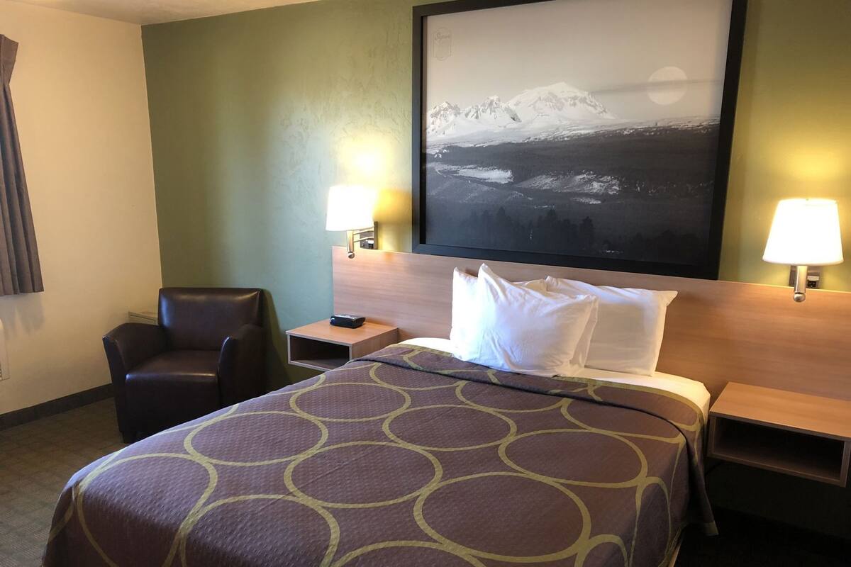 A hotel room with a bed, two nightstands, a chair, and a wall-mounted lamp.