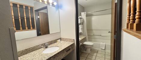 A bathroom with a granite countertop, a mirror with lights, a toilet, and a shower area.