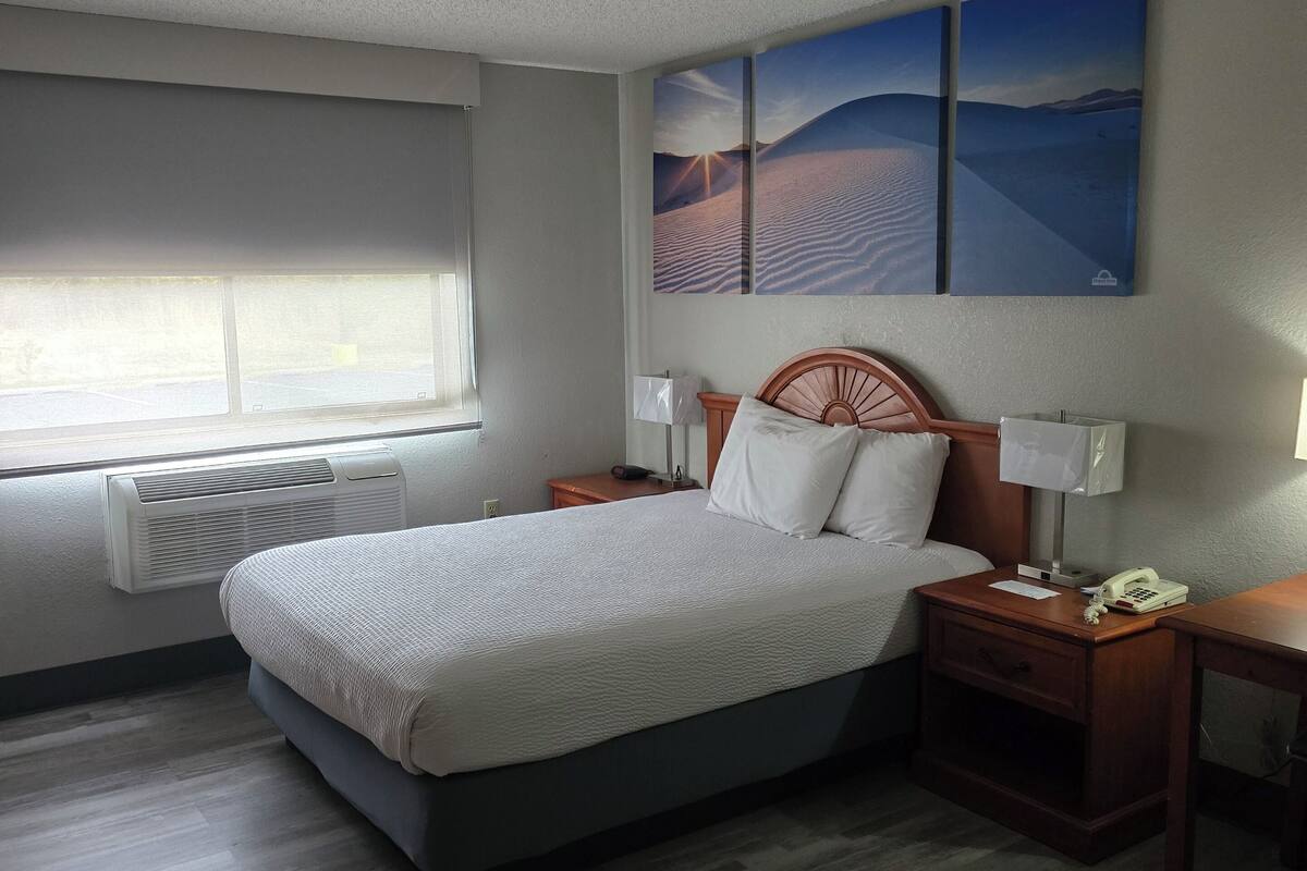 Hotelkamer met een bed, nachtkastje, bureau, raam met zonwering en een woestijnlandschap op de muur.