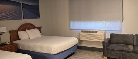 Een hotelkamer met een tweepersoonsbed, een bureau met stoel, een sofa, een raam met zonwering en een plafond met inbouwverlichting.