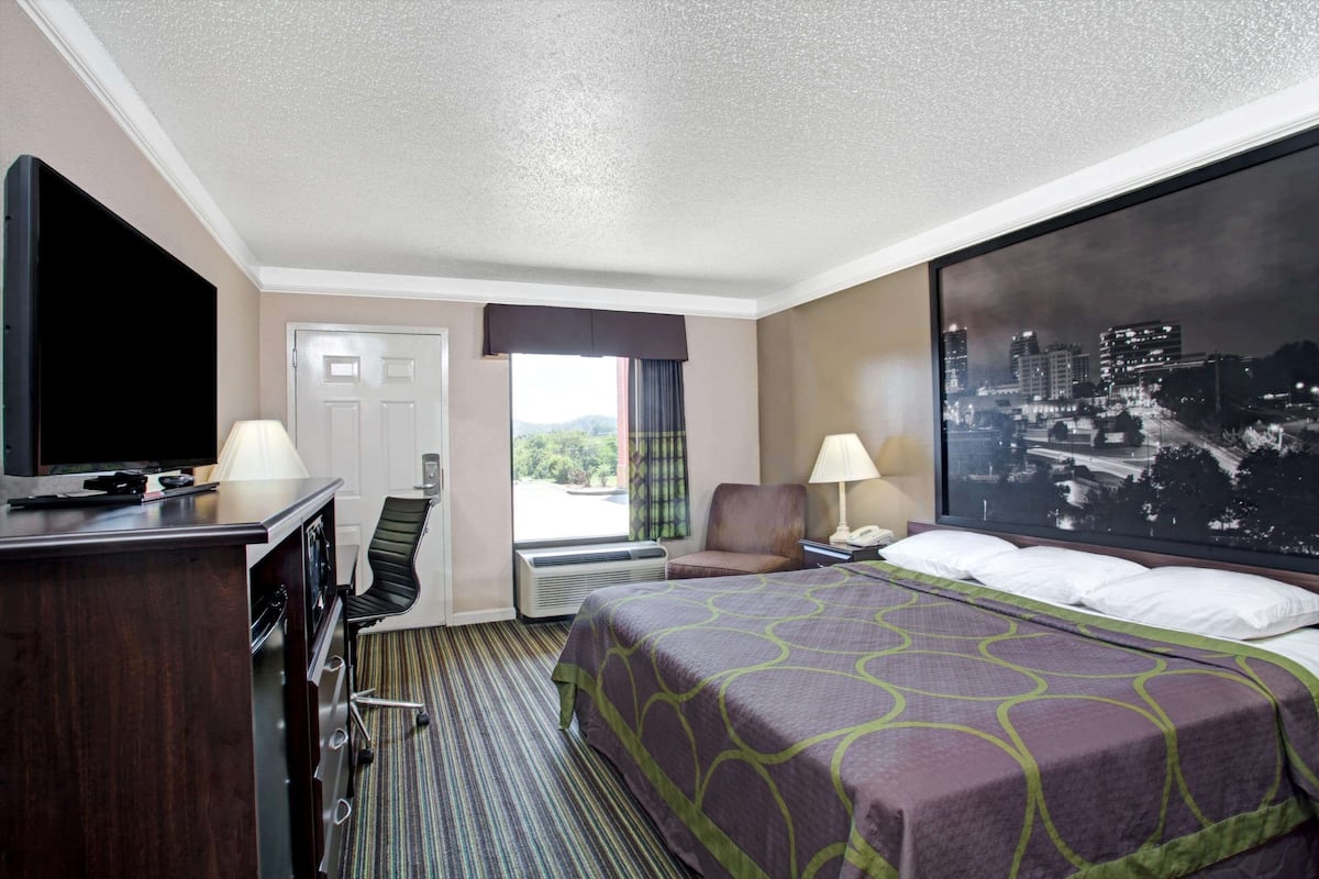 A hotel room with a bed, a desk with a TV, a chair, a window with a view, and a cityscape painting.