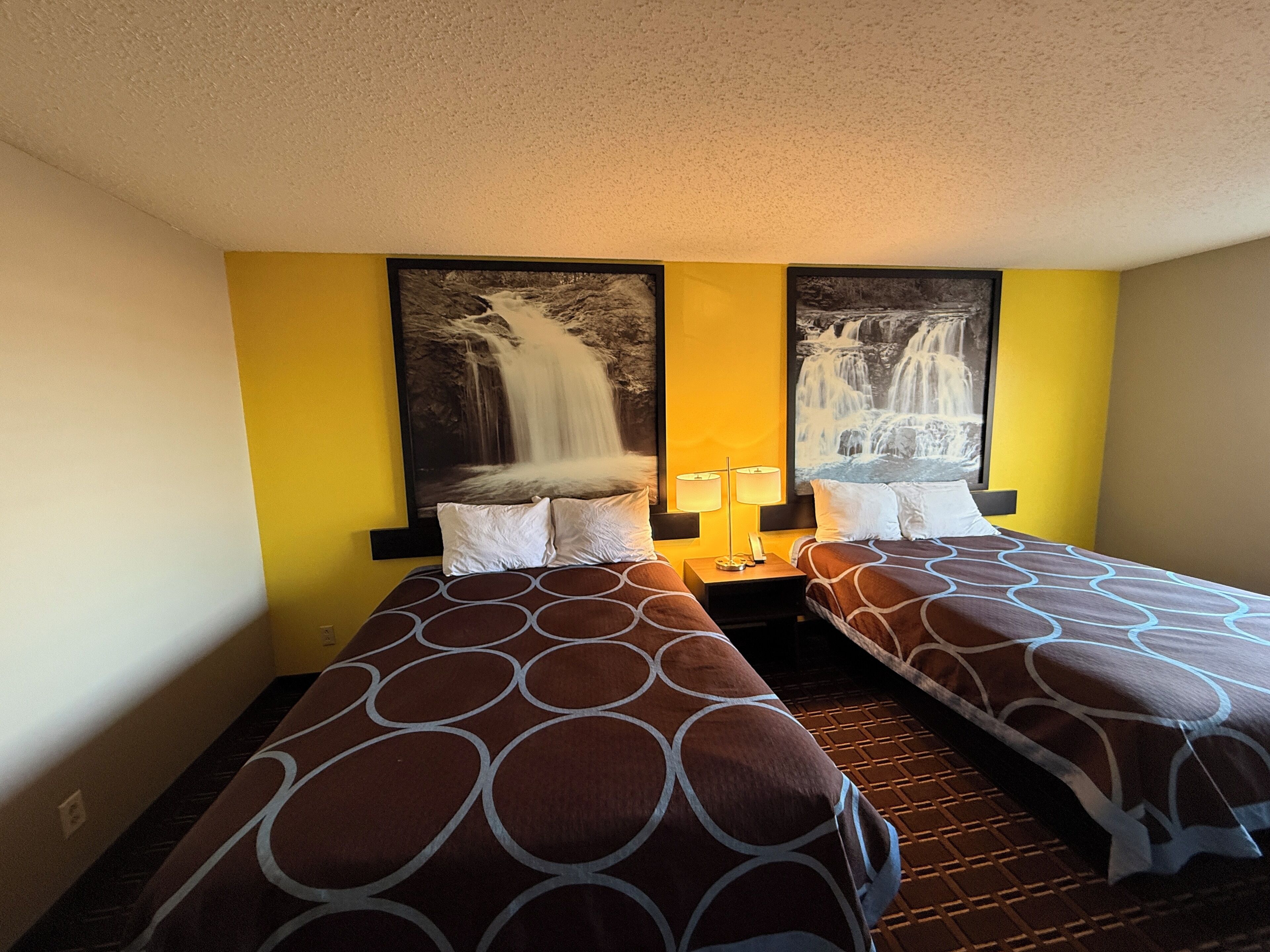 A hotel room with two beds, each with a patterned bedspread, a nightstand with a lamp, and two large wall-mounted paintings of waterfalls.
