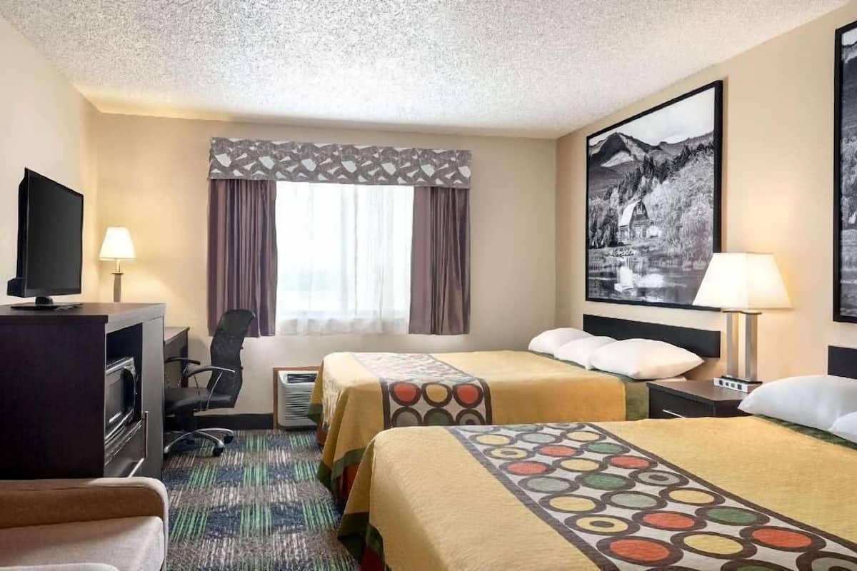 Habitación de hotel con dos camas, un escritorio con televisor, una silla, una ventana con cortinas y un cuadro en la pared.