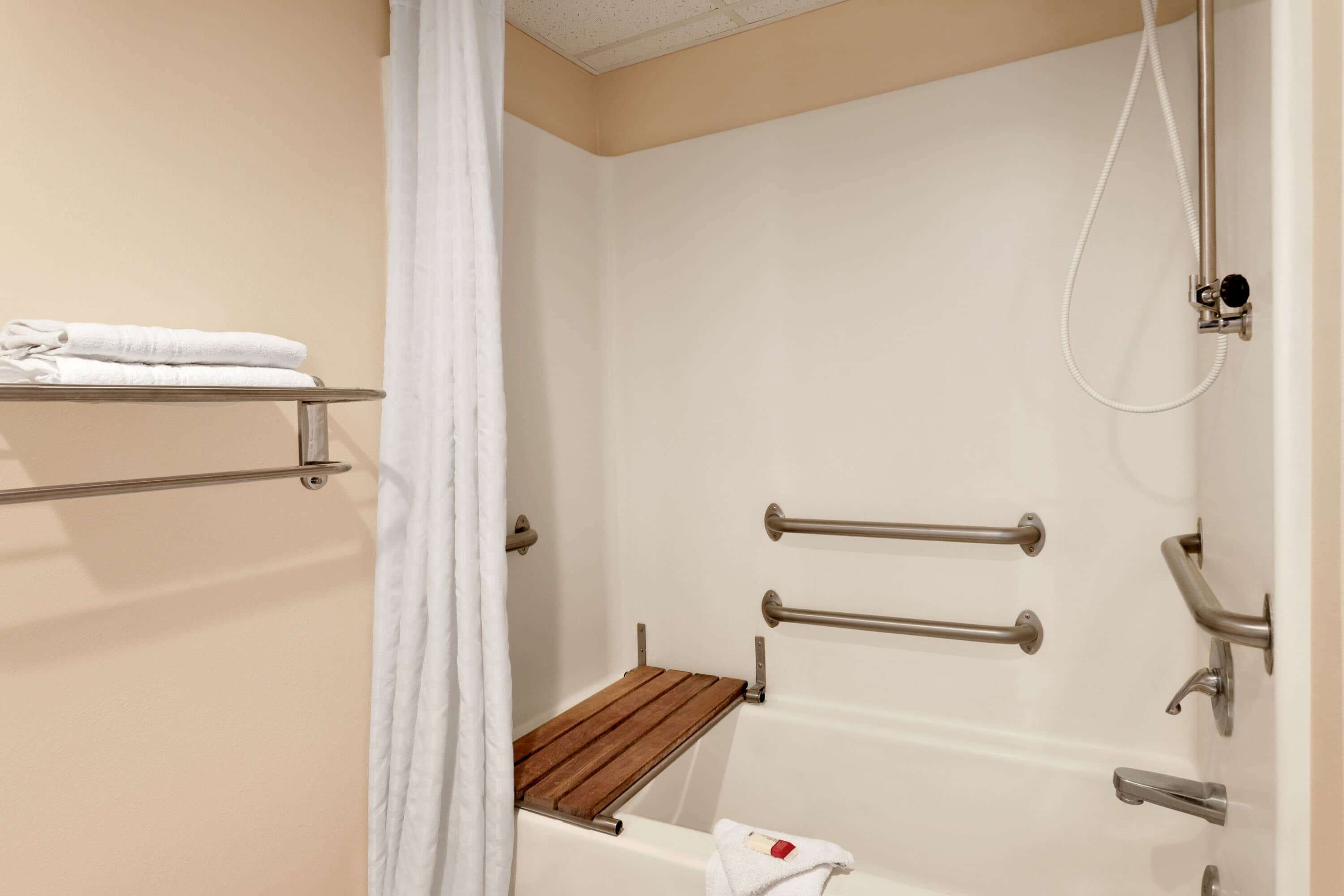 A bathroom with a shower, grab bars, a towel rack, and a wooden shelf.