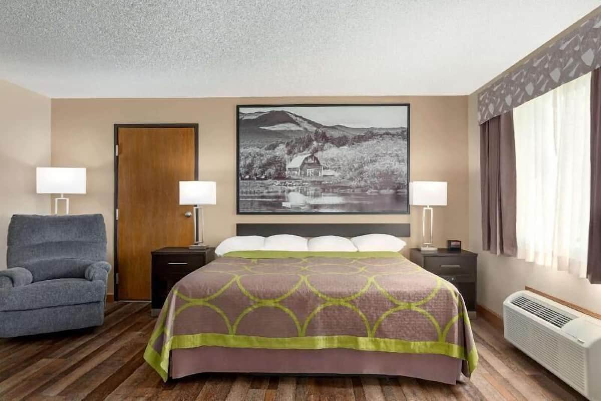 Habitación de hotel con cama, dos mesitas de noche, una silla, un cuadro grande en la pared y una ventana con cortinas.
