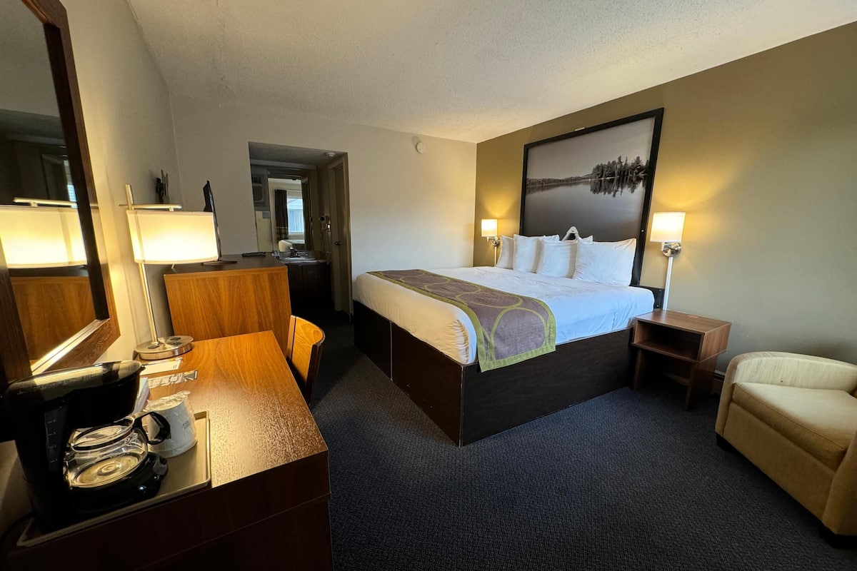 Una habitación de hotel con cama, lámparas de noche, un escritorio con cafetera, una silla y un cuadro en la pared.