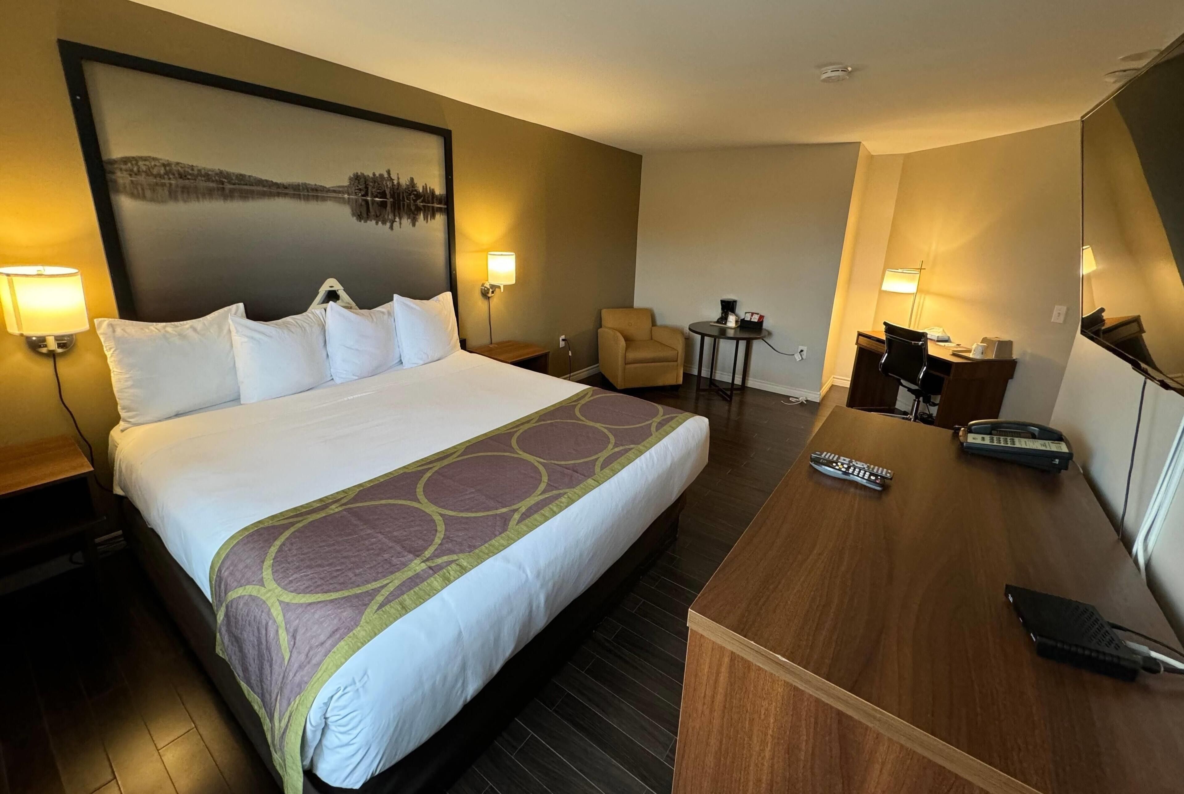 A hotel room with a large bed, a desk with a chair, a small table with a lamp, and a painting on the wall.