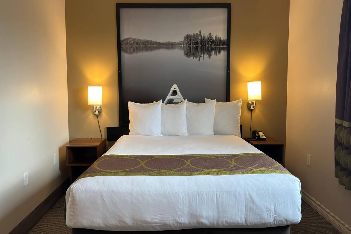 Habitación de hotel con una cama grande, dos lámparas de noche, una mesita de noche con un teléfono y un cuadro enmarcado de un lago.