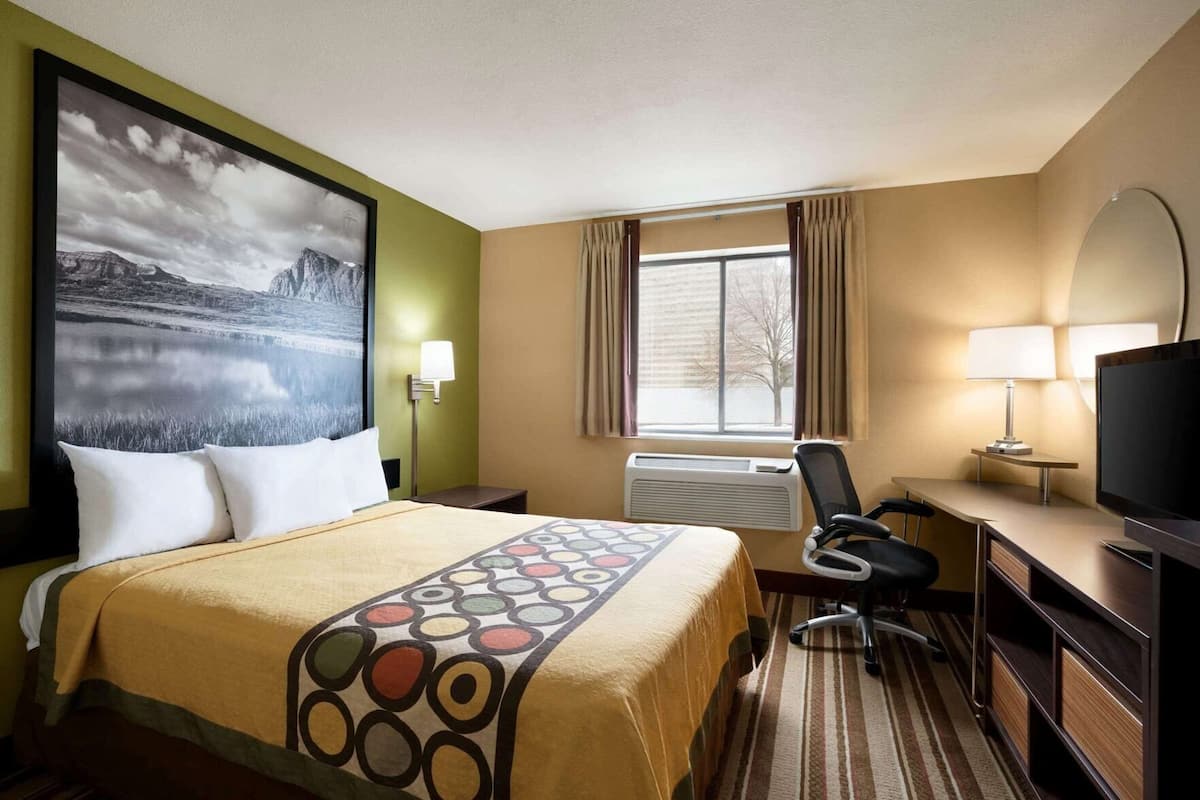 Une chambre d’hôtel comprenant un lit, un bureau avec une chaise, une télévision, une fenêtre avec des rideaux et une lampe fixée au mur.