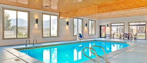 An indoor swimming pool with clear views of mountains through large windows, a wooden ceiling, and a poolside seating area.