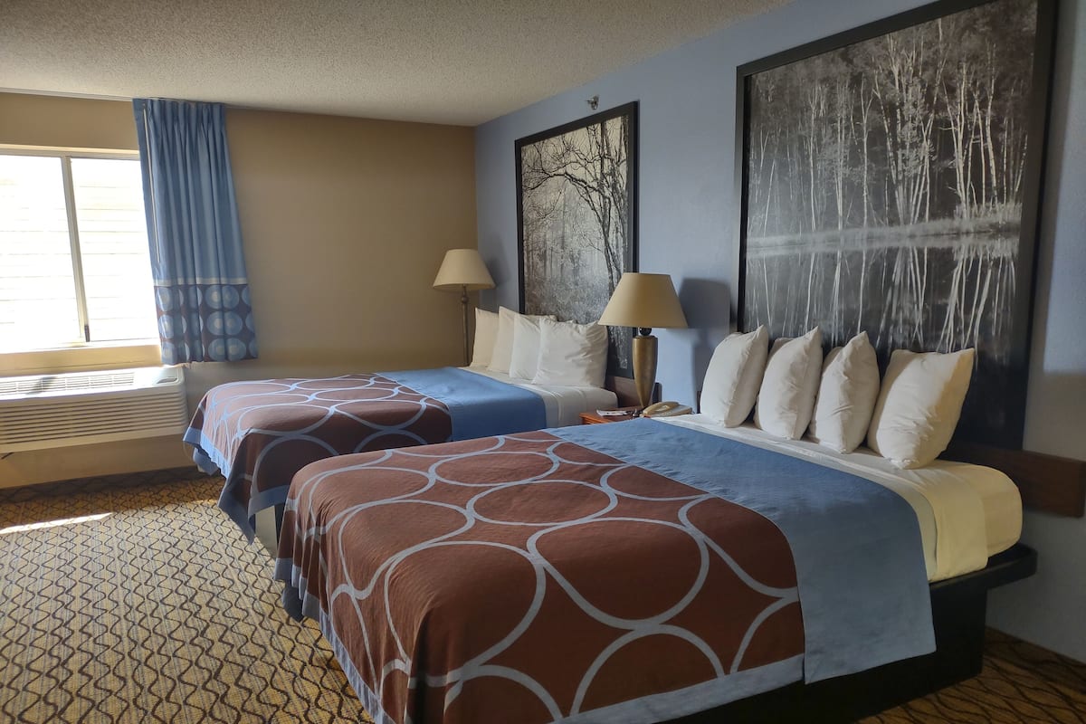 A hotel room with two beds, a large window with curtains, a wall-mounted lamp, and a painting.