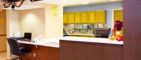 A reception area with a desk, computer, and a chair. There are yellow cabinets and a sink in the background.