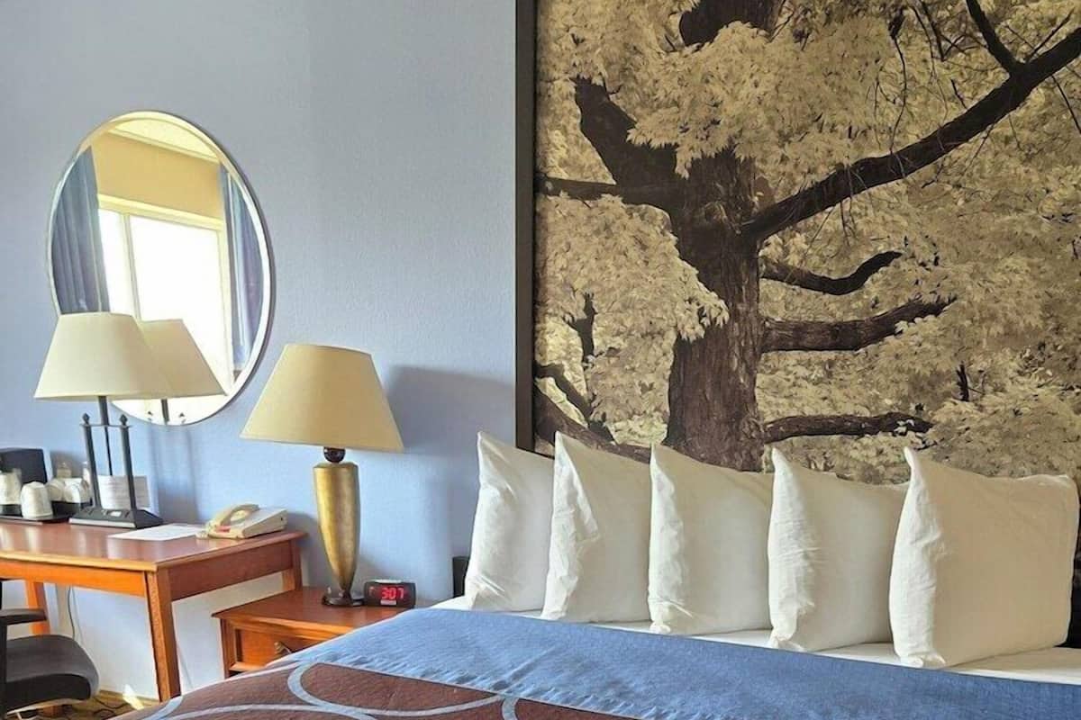 A hotel room with a bed, a desk with a lamp, a mirror, and a large wall art piece.