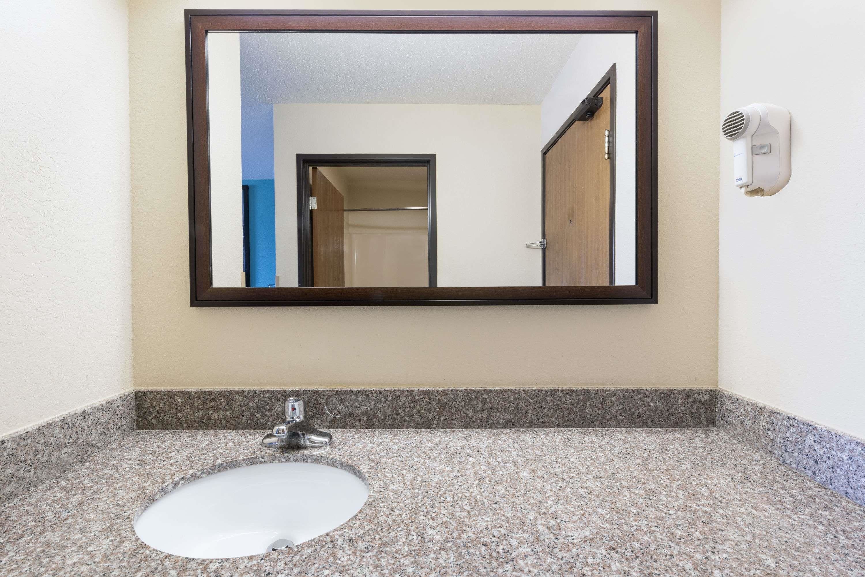 Baño con encimera de granito, lavamanos blanco, espejo y secador de pelo montado en la pared.