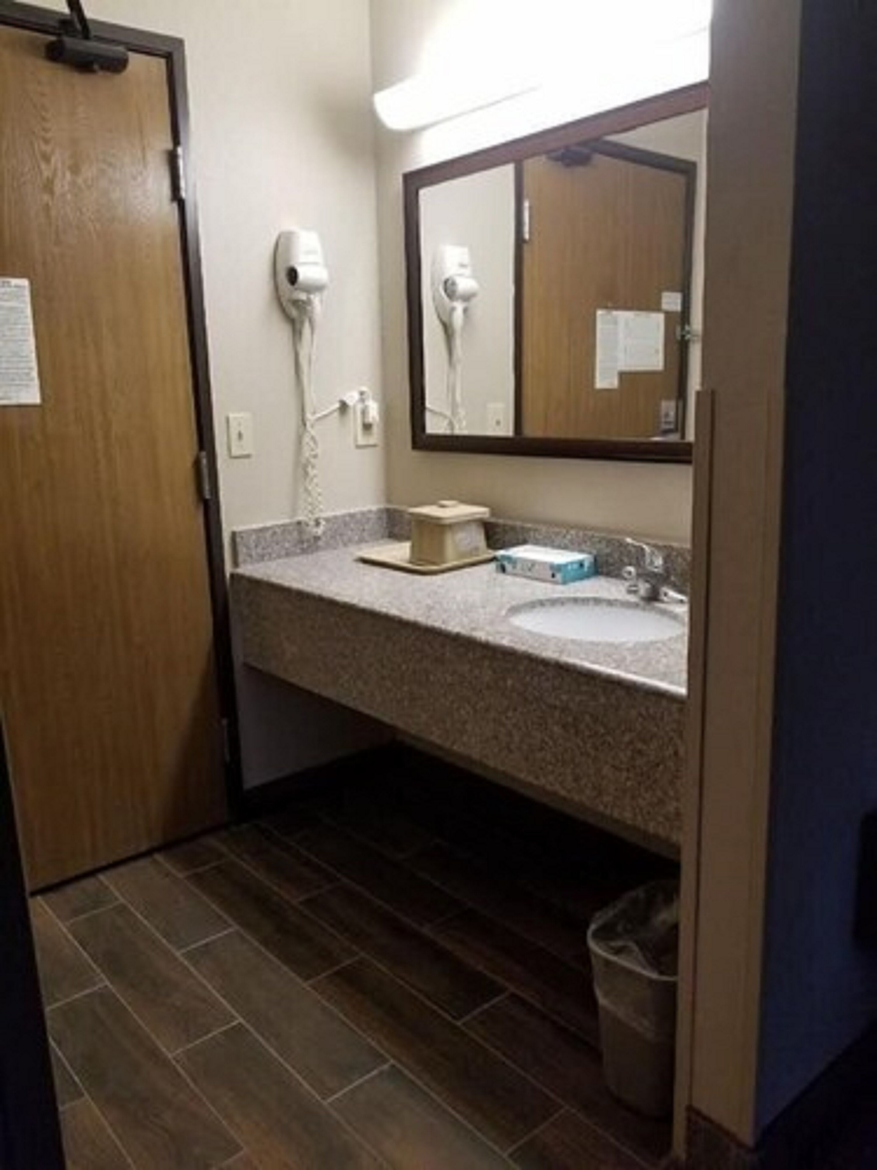 A bathroom with a wooden door, a mirror, a hairdryer, a sink, and a trash bin.