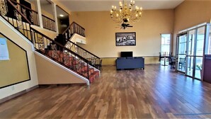 A spacious room with a wooden floor, a staircase with a patterned railing, a chandelier, and a large window.