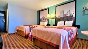 A hotel room with two beds, a small table, a chair, a mirror, and a cityscape picture on the wall.