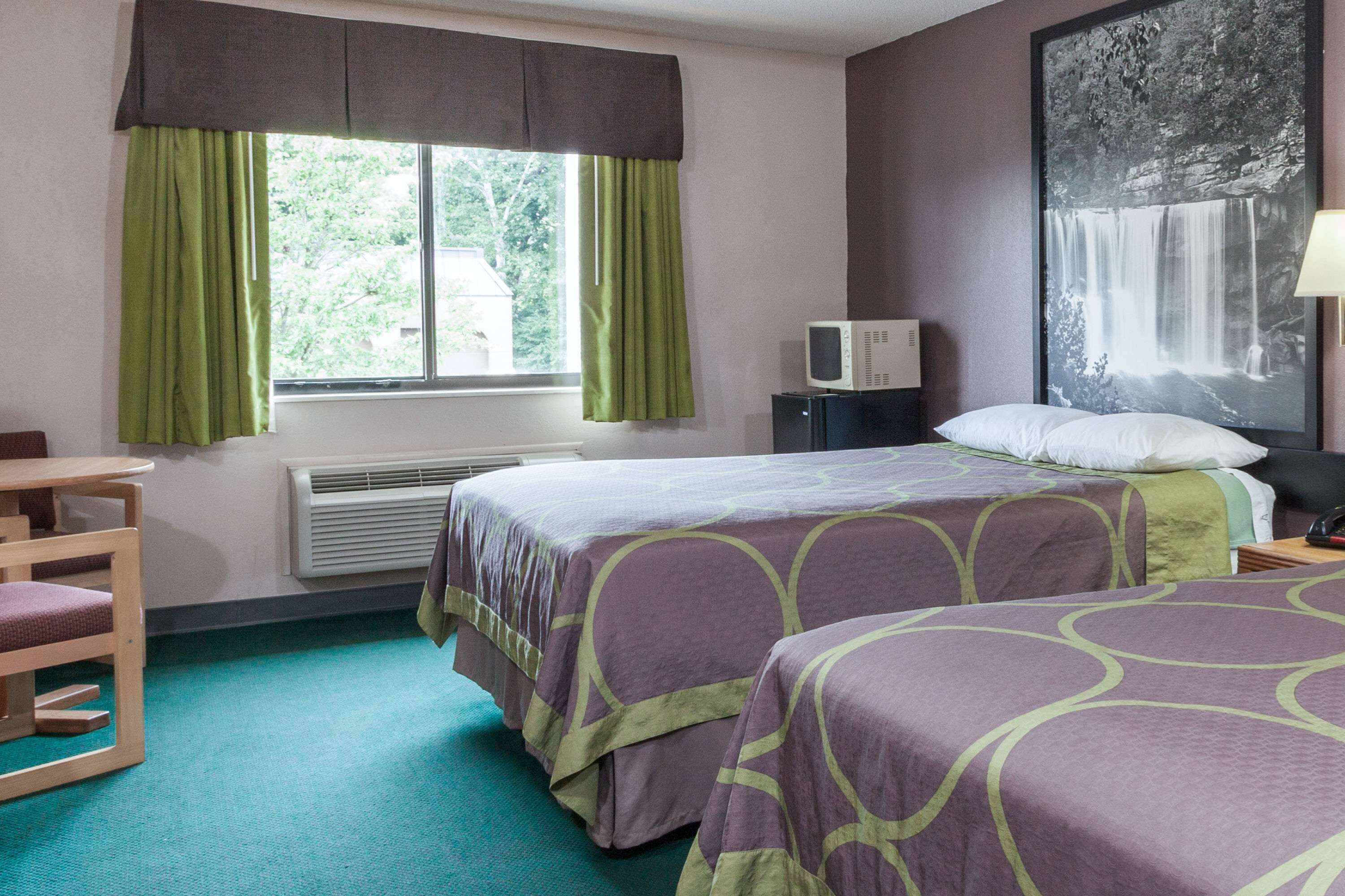 A hotel room with two beds, a desk, a chair, a window with curtains, a heater, a TV, and a wall-mounted lamp.