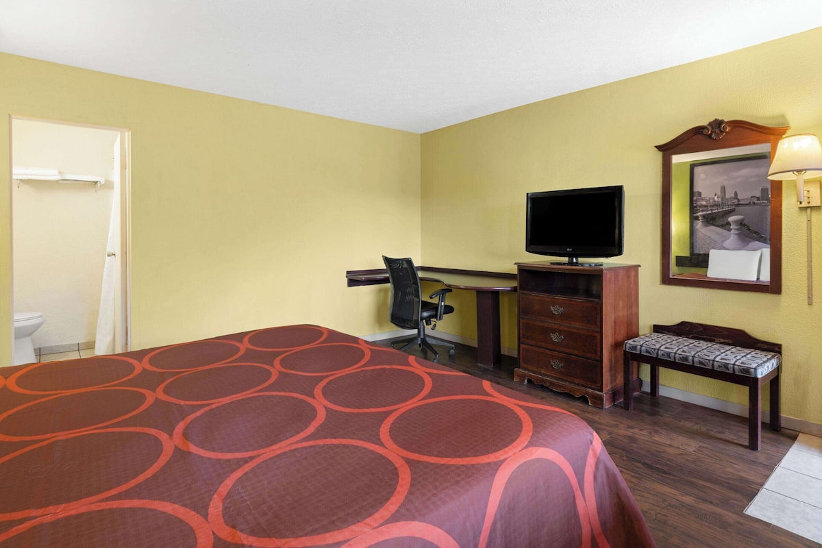 A hotel room with a bed, a desk with a computer, a television, a chair, a bench, and a mirror.