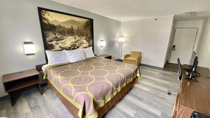 Quarto de hotel com uma cama grande, uma cadeira, uma escrivaninha com computador e um quadro na parede.