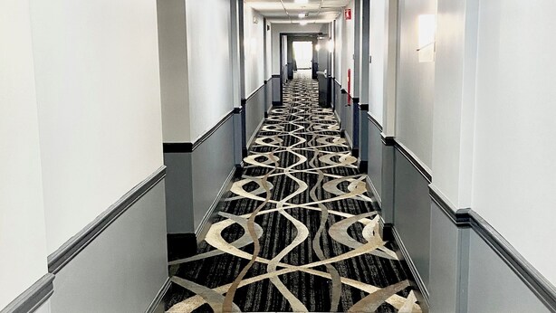 Um corredor de hotel com carpete estampado, paredes claras e chão de azulejos.