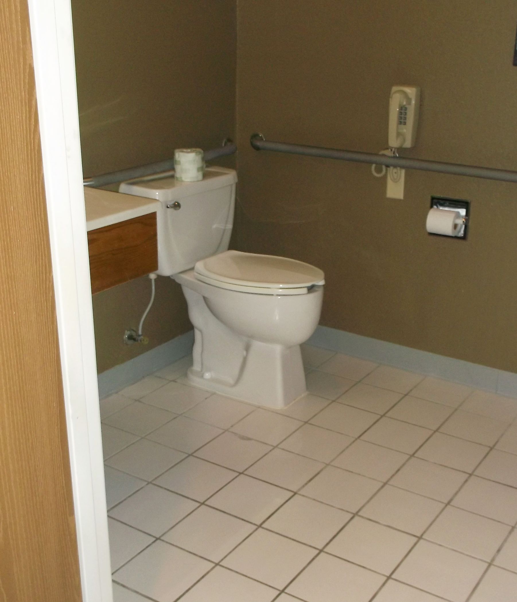 A small bathroom with a toilet, a wooden cabinet, and a handrail on the wall.