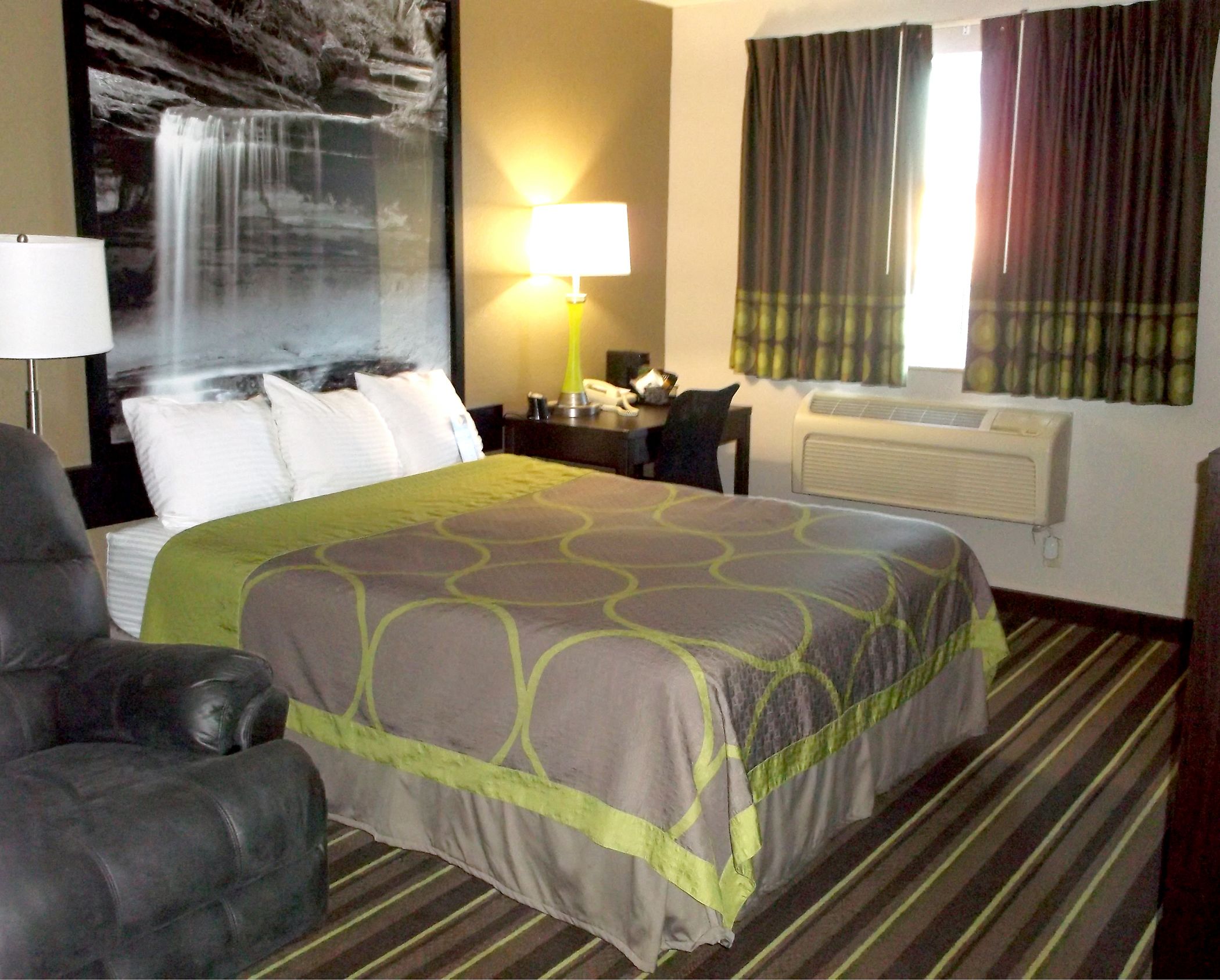 A hotel room with a large bed, a desk with a lamp, a chair, a window with curtains, and a wall-mounted air conditioner.