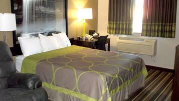 A hotel room with a large bed, a desk with a lamp, a chair, a window with curtains, and a wall-mounted air conditioner.