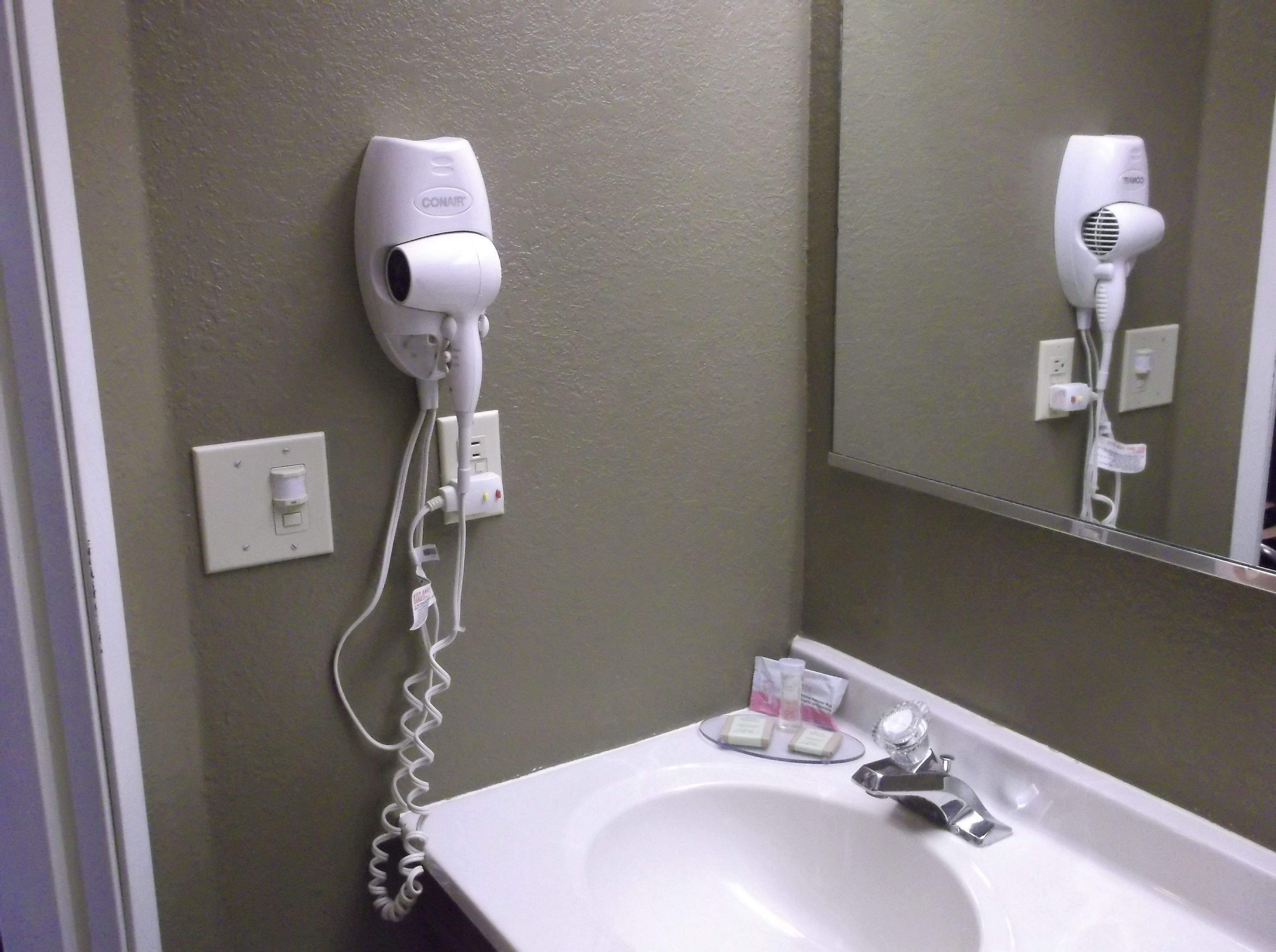 Une salle de bain avec un lavabo blanc, un sèche-cheveux fixé au mur et un miroir au-dessus du lavabo.