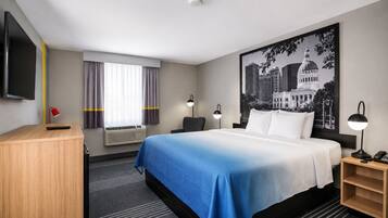A hotel room with a bed, a desk, a TV, a window with curtains, a wall-mounted lamp, and a large cityscape poster.