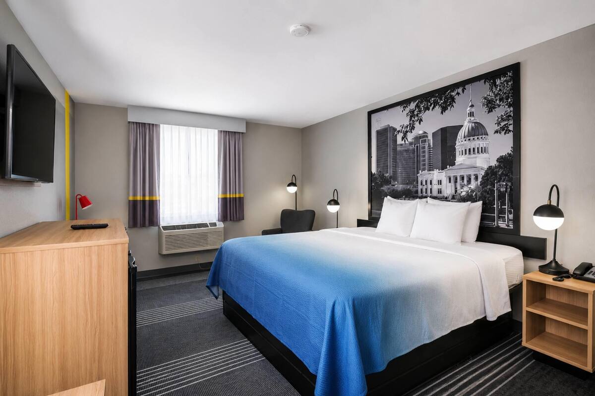 Una habitación de hotel con cama, escritorio, televisor, ventana con cortinas, aire acondicionado de pared y un cuadro enmarcado.