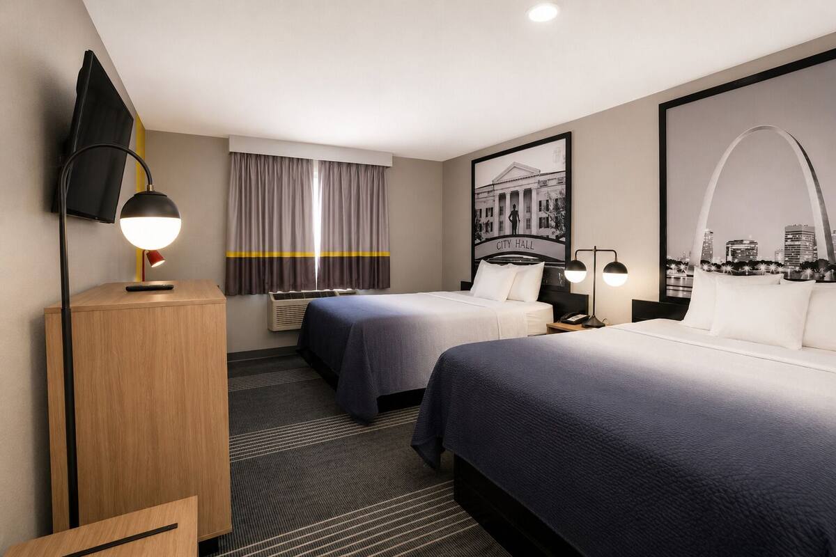 Habitación de hotel con dos camas, un escritorio, un televisor de pantalla plana, un ventanal con cortinas y vistas a la ciudad.