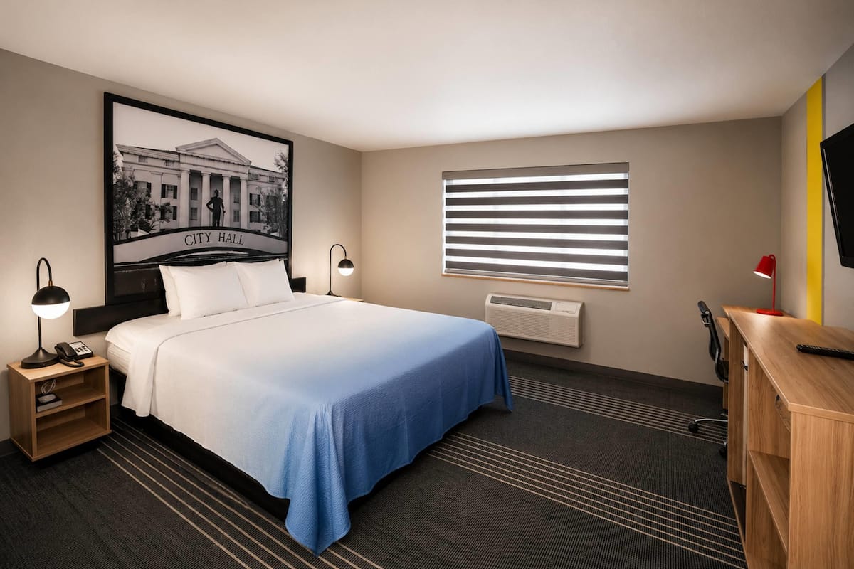 Una habitación de hotel con cama, mesita de noche con lámpara, escritorio, silla, un cuadro con una imagen de un edificio, ventana con persianas y televisor montado en la pared.