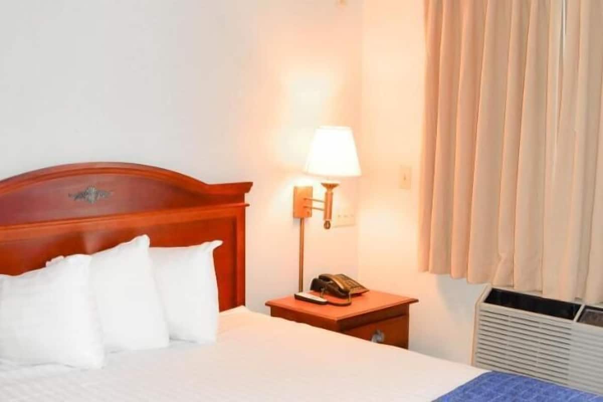 A hotel room with a wooden headboard, a bedside lamp, a telephone, a heater, and a window with blinds.